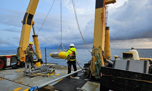 Two people wearing safety vests and helmets work on the deck of an offshore platform, surrounded by open ocean. A large object is hanging from cables near the edge of the platform.