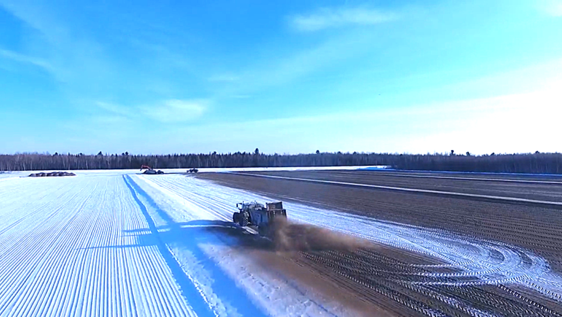 Wide shot of a farmer spreading sand on a flat, snowy surface.