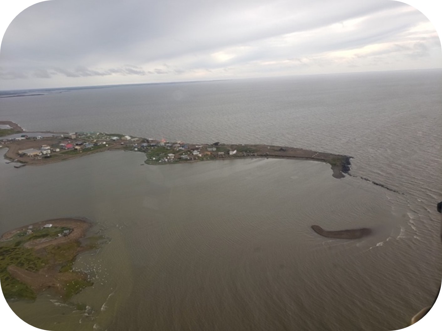 Aerial view of a small coastal community on a narrow peninsula surrounded by water under overcast skies.