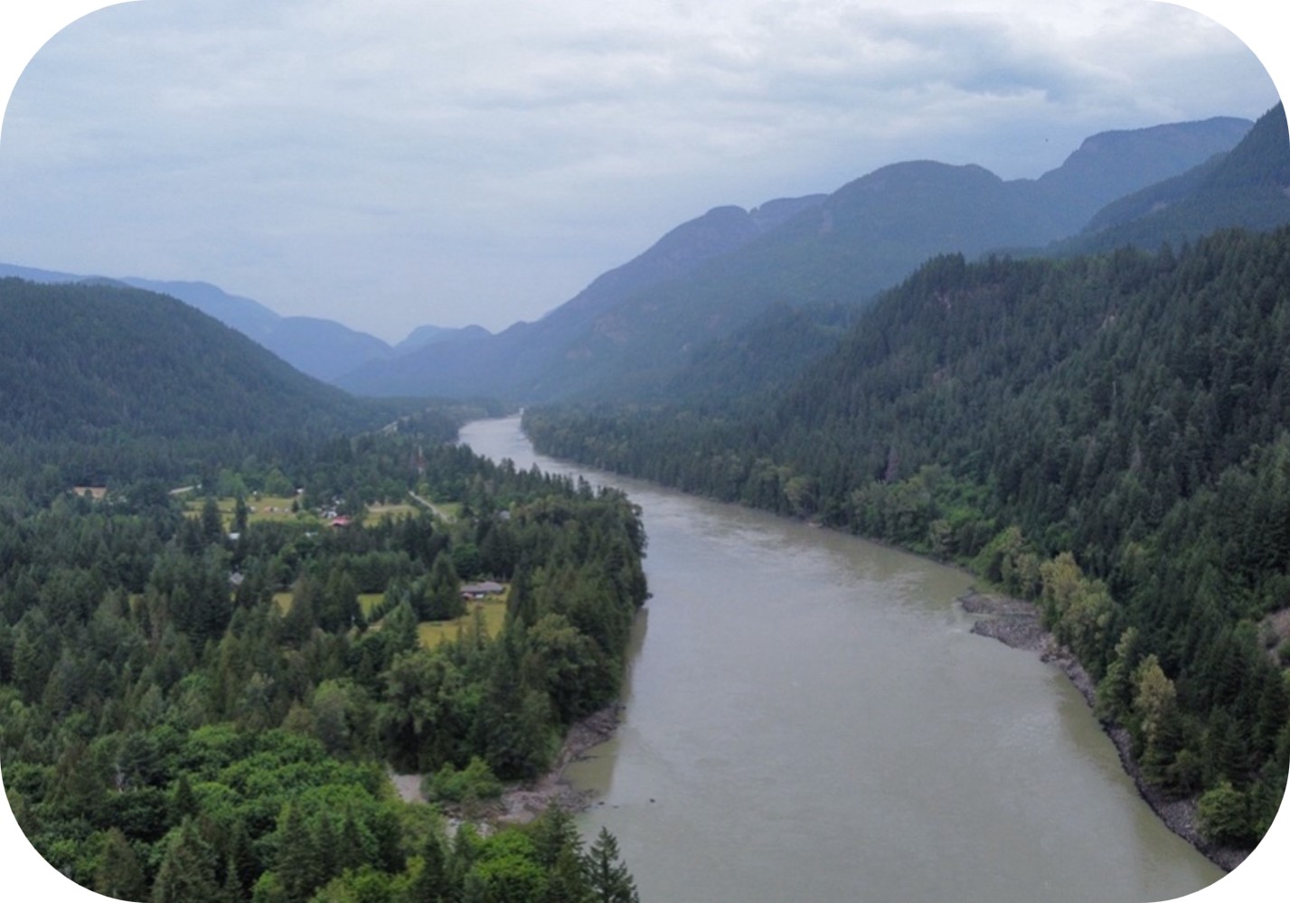 Aerial view of a river flowing through a forested mountain valley with rural buildings under a cloudy sky.