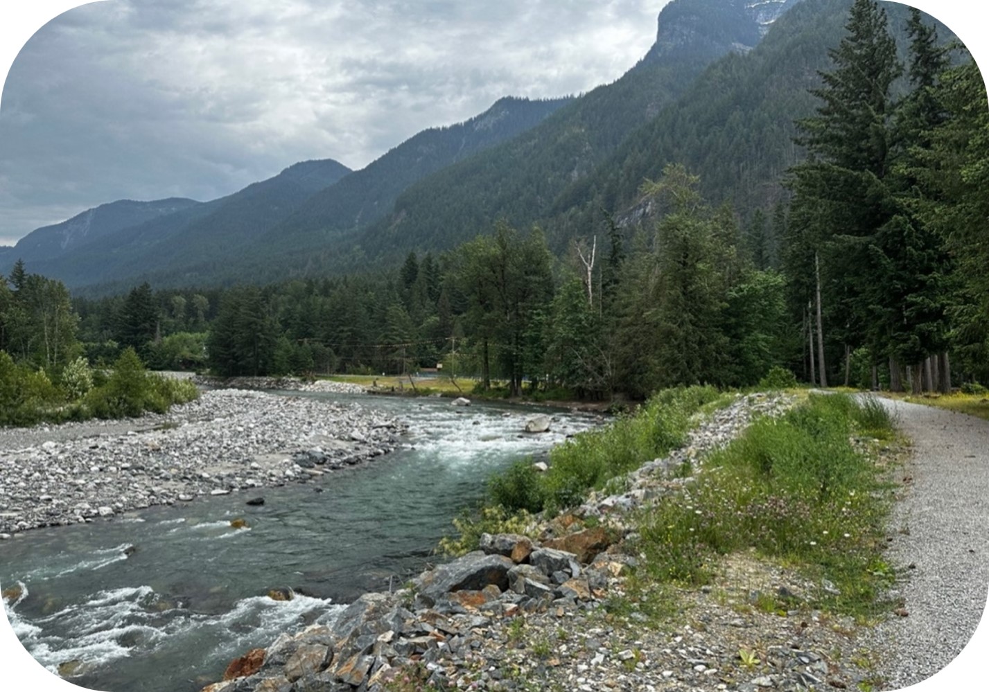River through a forested mountain valley, bordered by a gravel trail under an overcast sky.
