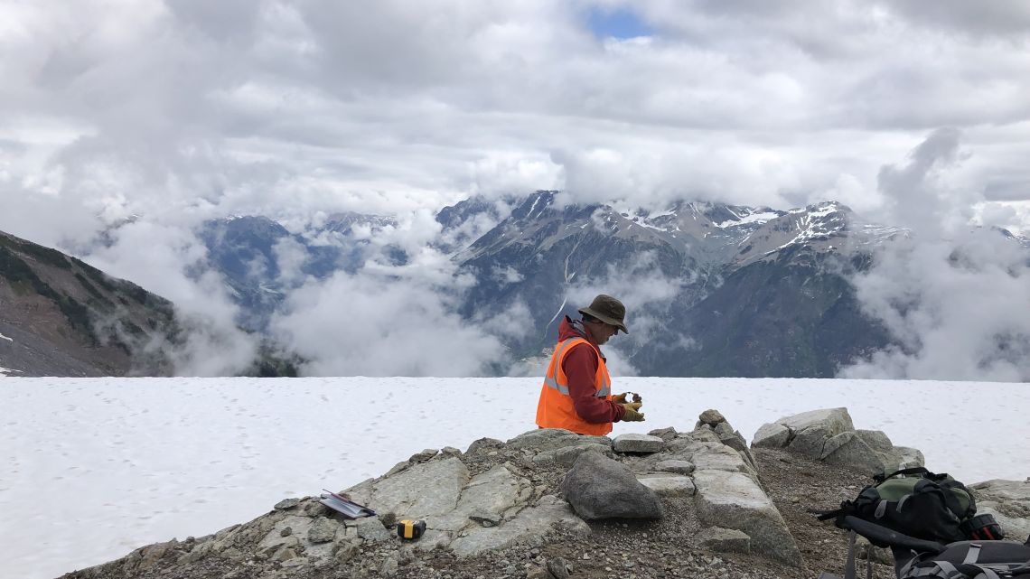 Researcher in an orange safety vest examines a rock sample on a rocky, snow-covered mountainside, with cloud-covered mountains in the background.