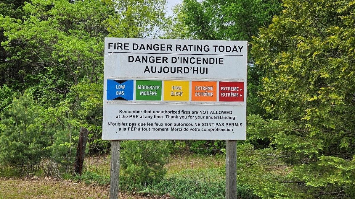 A bilingual fire danger rating sign in a wooded area displays a colour-coded scale ranging from low to extreme plus.