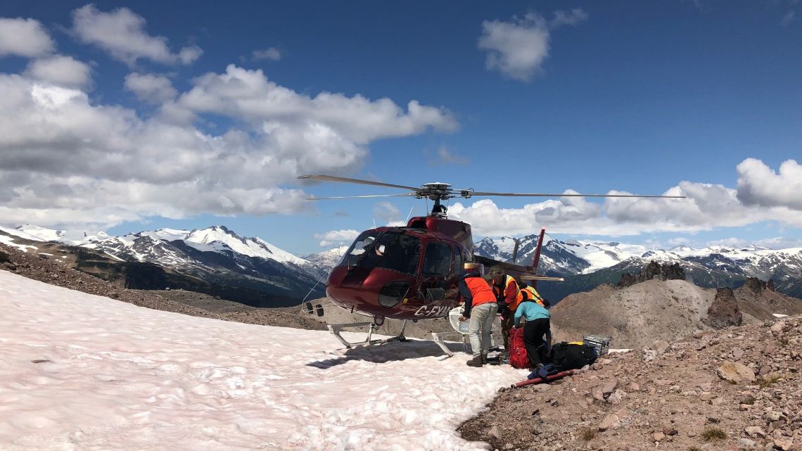 Researchers load gear on to a red helicopter on a snowy mountainside, with rugged, snow-capped peaks in the distance.