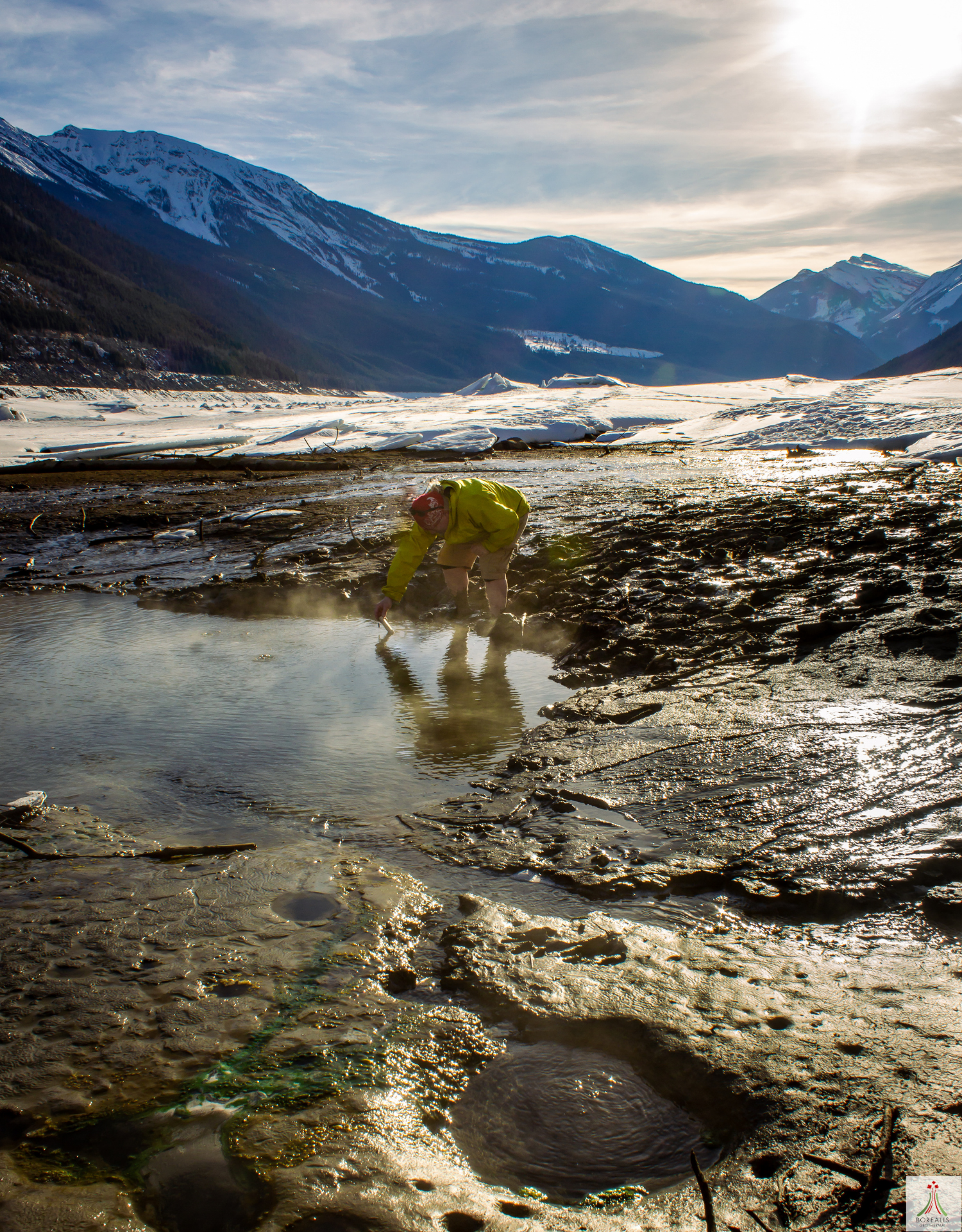 A picture containing a single person in a yellow-green jacket reaching down to get a sample of steaming water from a hot spring pool. Around the hot water pool is mud and a rock foundation that has holes with hot water. Behind the dirt, snow goes up to the tree-filled mountains. The sun shines on the camera, and the clouds appear like wrinkles in the sky.