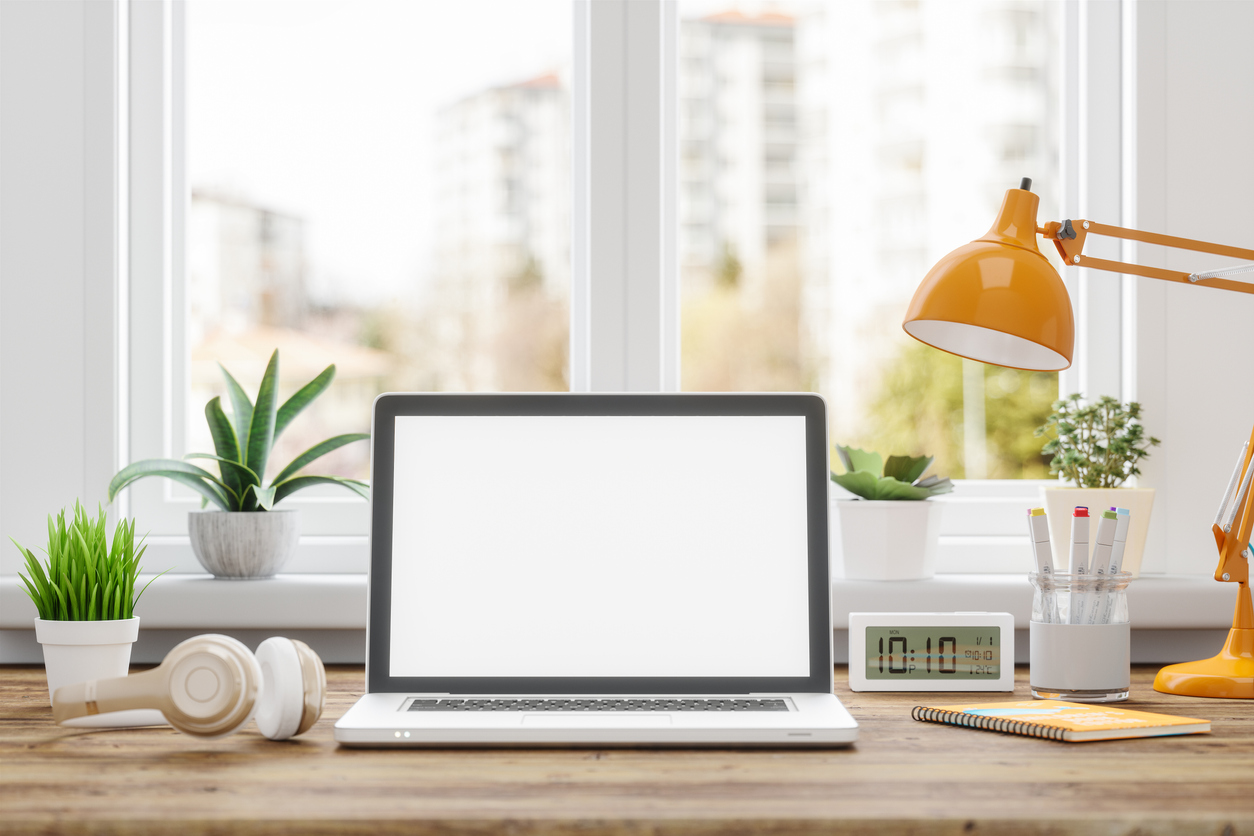 Desk with office supplies such as desk lamp, note pad, pens and earphones, as well as a laptop with a blank screen.