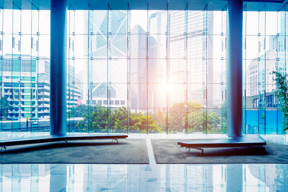 This image shows a view from inside a modern building, looking out through a large glass wall with the sun shining in. Other buildings can be seen in the distance.