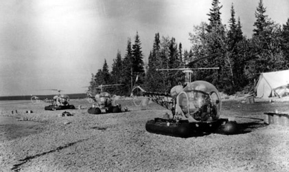 Three helicopters on the ground with a tent to the left side with trees in the background
