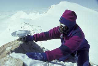 Technician on snowy mountaintop in winter attire holding a GPS antenna with a mountainous backdrop