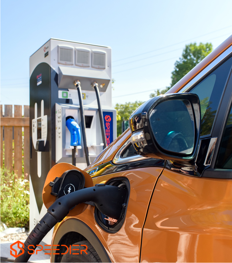 An electric vehicle charger, situated in a parking lot in front of a fence.