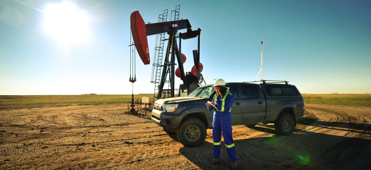 Flux Lab Researcher, Jennifer Baillie, with research truck parked in front of oil field pump jack located in Weyburn, SK. Jennifer takes notes on a clipboard outside of the research truck. 