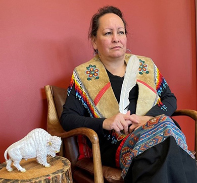 Elder Heather Poitras. She is wearing a black shirt, a black skirt with a blue patterned stripe, and an animal hide vest with beaded tassels. 