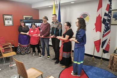 A group of six people stand in a line holding a red ribbon that is about to be cut. Beneath their feet is a circular Medicine Wheel rug.