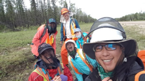 A selfie of six smiling people in high-visibility vests and rain jackets wearing bug nets over their heads. The background is a field of short grass and a stand of pine trees.