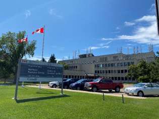 A large concrete building against a bright blue sky with two Canadian flags flying in front. Trucks and cars are parked in a lot in front of the building. A sign reads “Northern Forestry Centre / Centre de foresterie du Nord.”