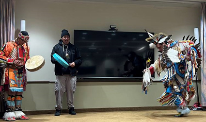 Three men are performing on stage. Two are wearing cultural regalia: bright orange and blue garments adorned with feathers, tassels, furs, and beads. One man is dancing while holding an eagle feather fan. The two others are drumming on handheld drums.