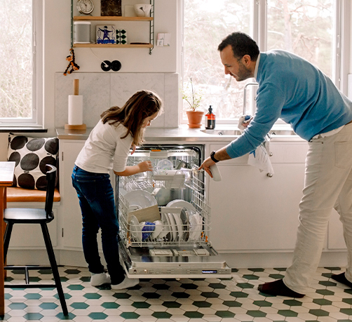 Parent and child arranging utensils in dishwasher while standing at kitchen.