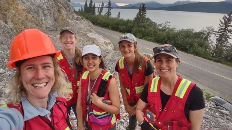 Group of five women wearing high visibility vests on a rocky ledge near a lake.