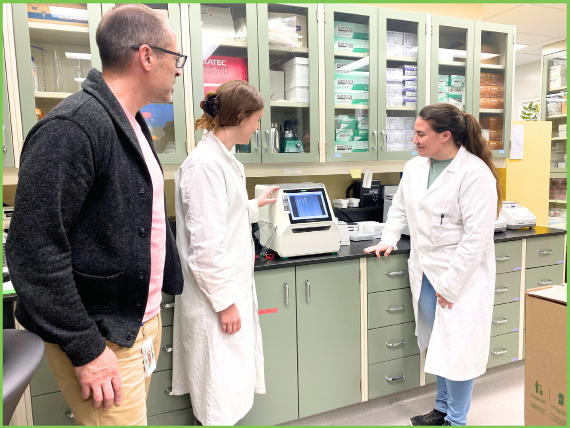 Man in sweater and two women in lab coats look at a machine that analyzes DNA