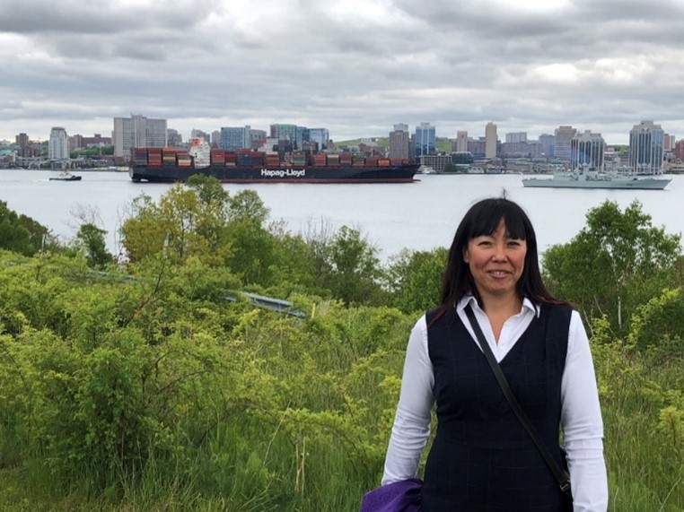 Woman standing on grassy hill with body of water, barges and Halifax skyline in the background.