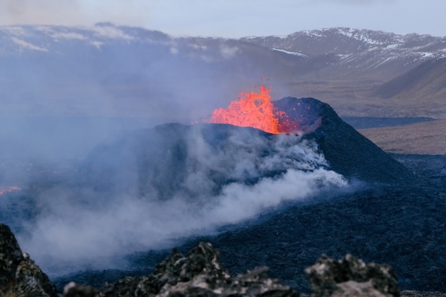 Photo showing molten lava spewing from a volcano against the backdrop of a rugged mountainous landscape.