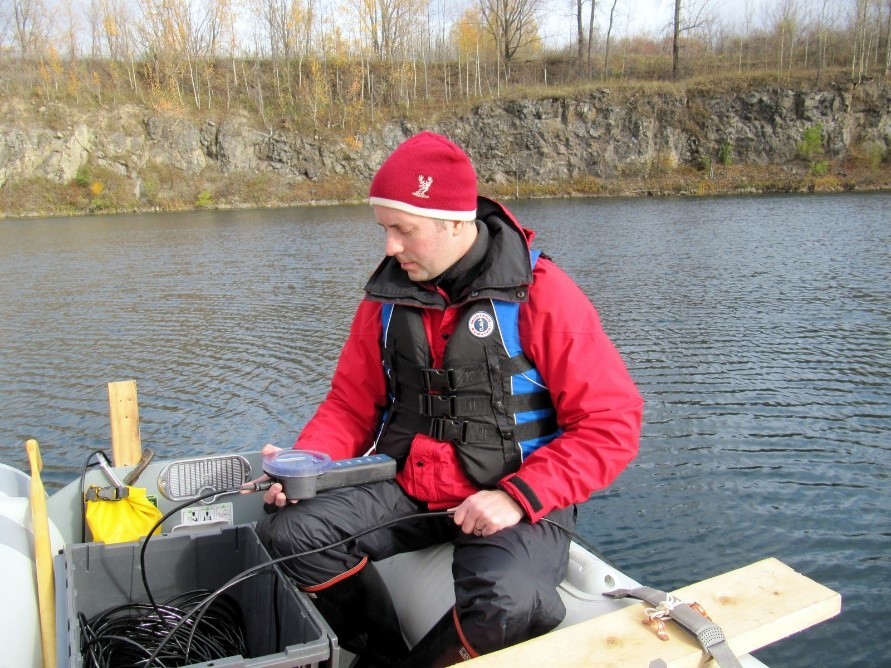 Michael Parsons sits on a small boat in the middle of a body of water with a measuring device in his hand to determine water quality at the former mine in Oka, QC.