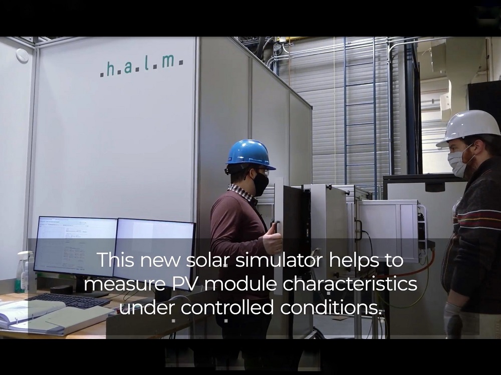 Two men in hard hats outside of solar simulator in lab setting. On screen text: This new solar simulator helps to measure PV module characteristics under controlled conditions